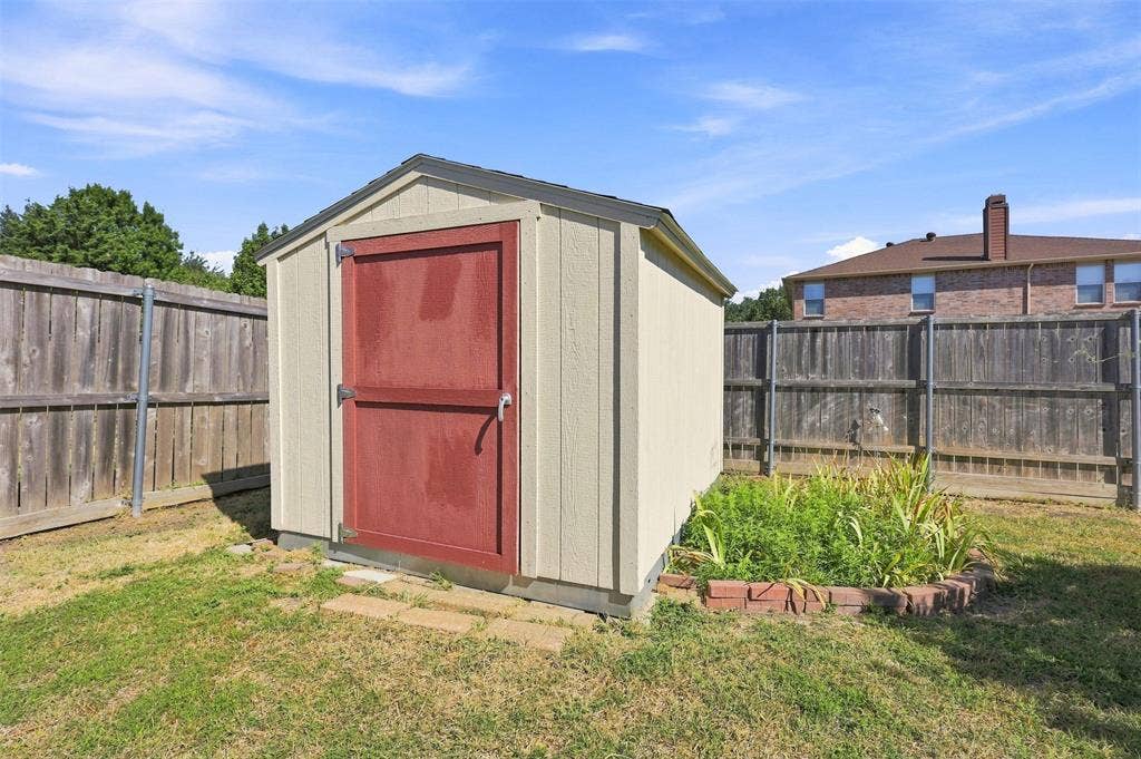 1406 Quail Meadow Drive, Wylie, TX 75098 Wood-paneled storage shed with a red door, featuring a shingled roof, rain gutter, and a surrounding wood fence