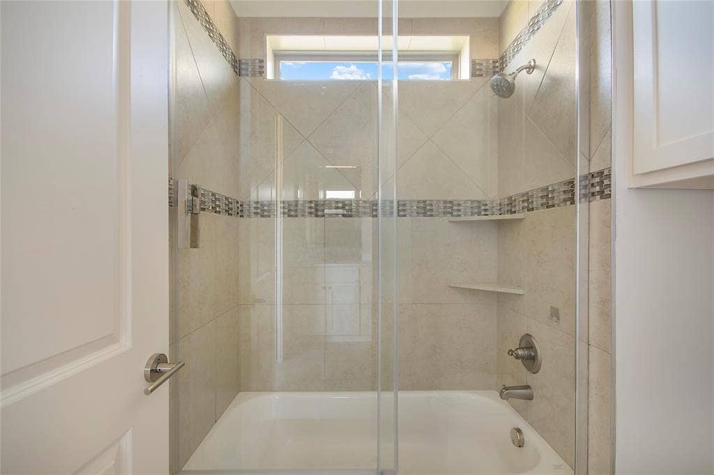 Bathtub and shower combination featuring a glass enclosure, tiled surround with decorative accent band, and built-in shelving