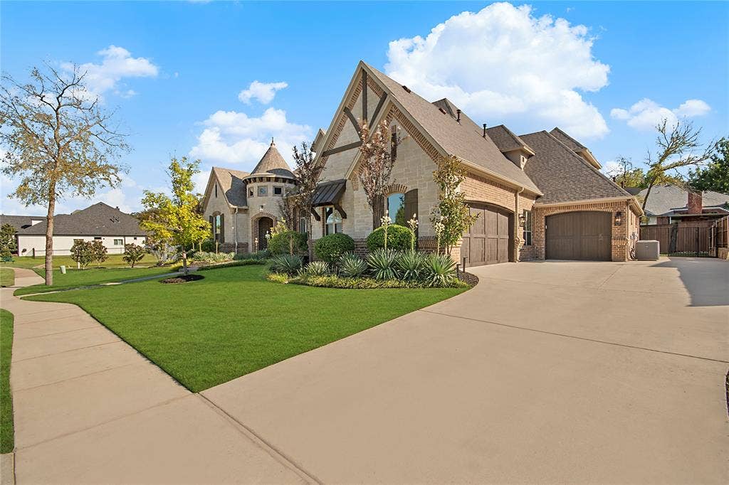 Stone and brick facade with a prominent turret, multiple gables, a well-maintained lawn, and a wide paved driveway leading to a three-car garage