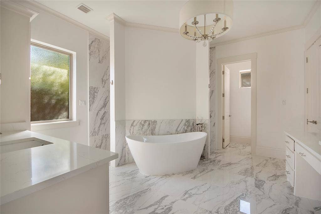 Freestanding soaking tub, white vanity with integrated sink, marble-look tile flooring, and a decorative chandelier