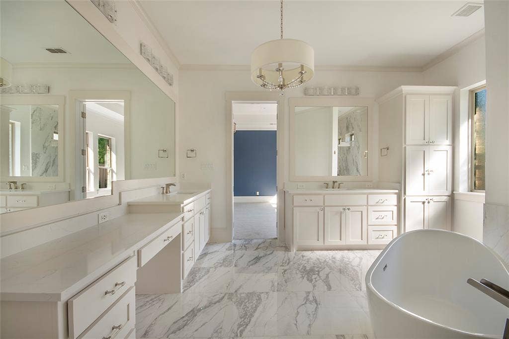 Freestanding soaking tub, dual vanities with white cabinetry, light-toned countertops, and marble-look flooring