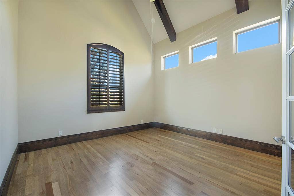 Vaulted ceiling with exposed wood beams, wood-finish flooring, dark wood baseboards, and arched window with plantation shutters
