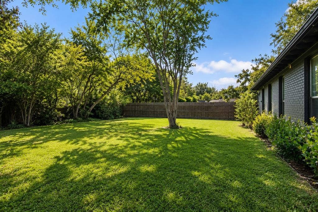 5012 Wateka Drive, Dallas, TX 75209 Expansive lawn with mature trees and a privacy fence, complemented by a brick exterior and garden beds