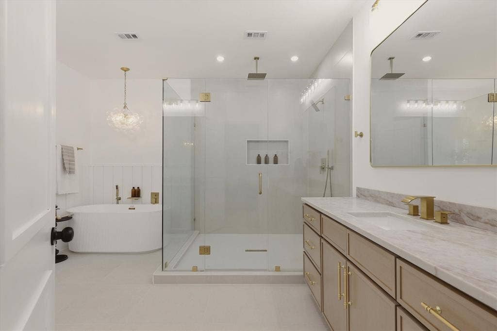 Freestanding soaking tub, frameless glass shower enclosure with dual showerheads, dual vanity with undermount sinks, stone countertop, and gold-finish hardware throughout