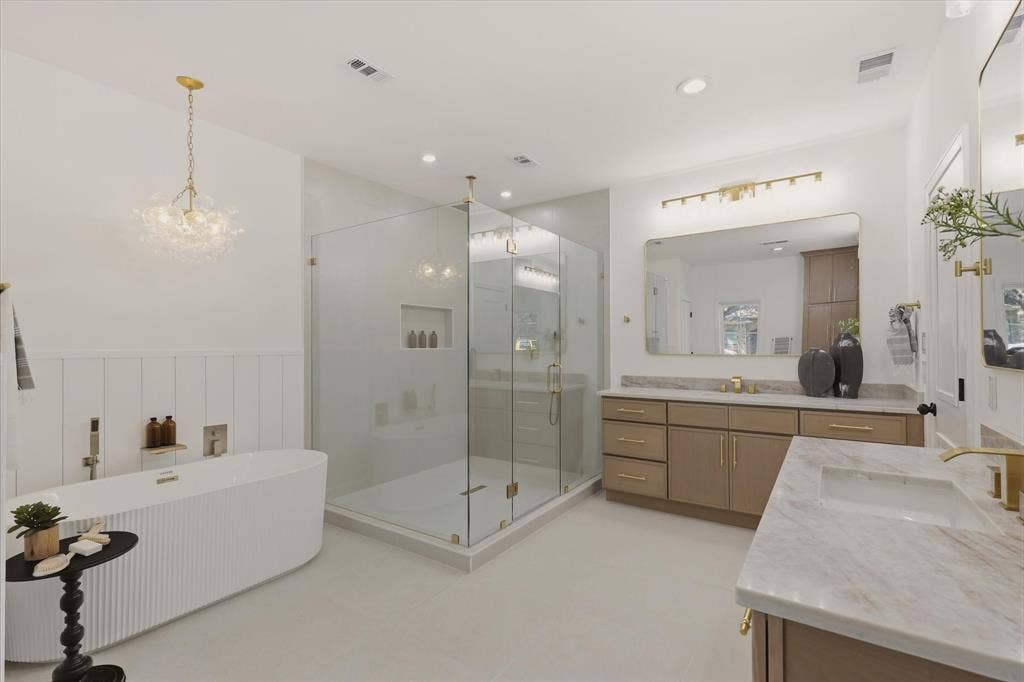 Freestanding soaking tub with vertical fluting, a spacious glass-enclosed shower, dual vanity with undermount sinks, stone countertops, and gold-toned hardware