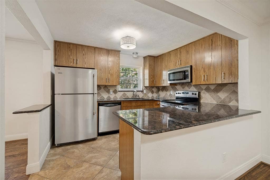 5818 E University Boulevard, #138, Dallas, TX 75206 Kitchen featuring wood-finish cabinetry, stainless steel appliances, dark stone countertops, and a patterned tile backsplash