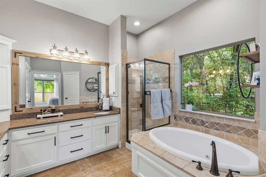 6810 Trinity Falls Parkway, McKinney, TX 75071 Dual vanity with white cabinetry and dark hardware, framed mirror with integrated lighting, tiled flooring, built-in soaking tub with dark fixtures, and a glass-enclosed shower