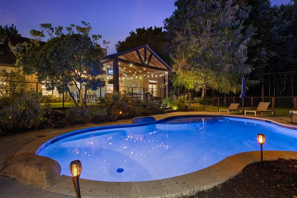6810 Trinity Falls Parkway, McKinney, TX 75071 Curved swimming pool with stone coping, a dedicated outdoor living structure, mature trees, and perimeter fencing