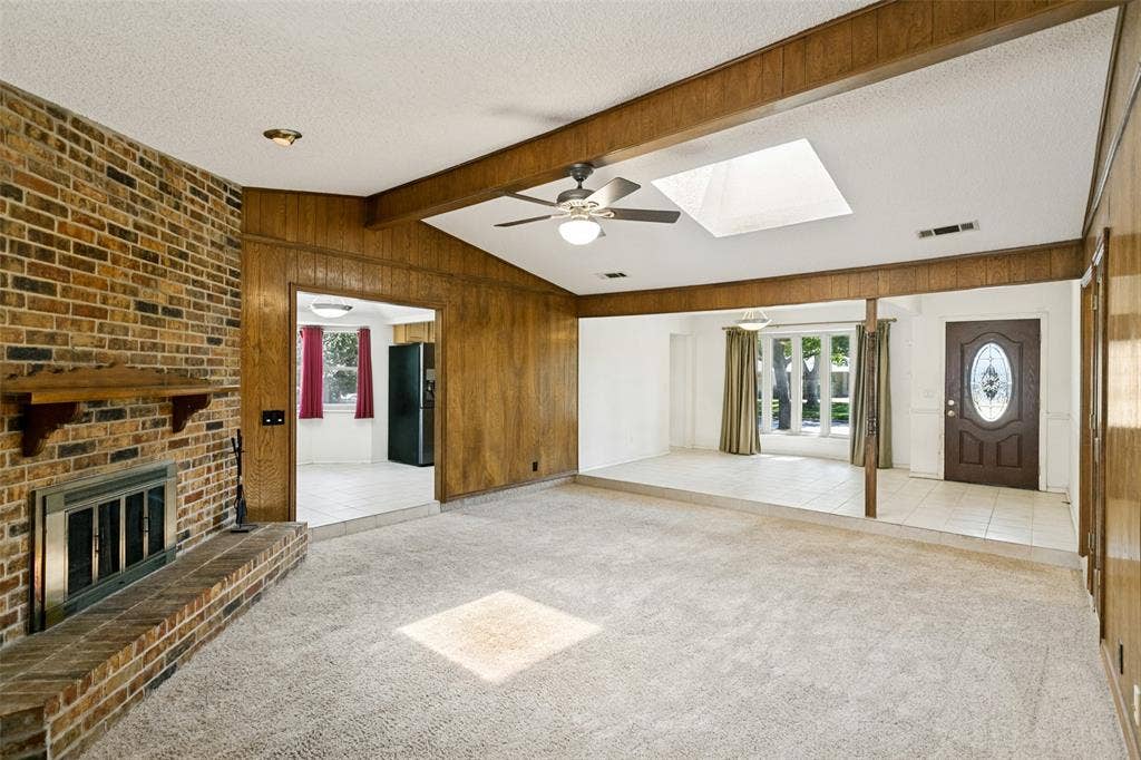 2717 Pinehurst Drive, Plano, TX 75075 Vaulted ceiling with a skylight and ceiling fan, exposed brick fireplace with a hearth and wood mantel, wood paneling, and a decorative front entry door