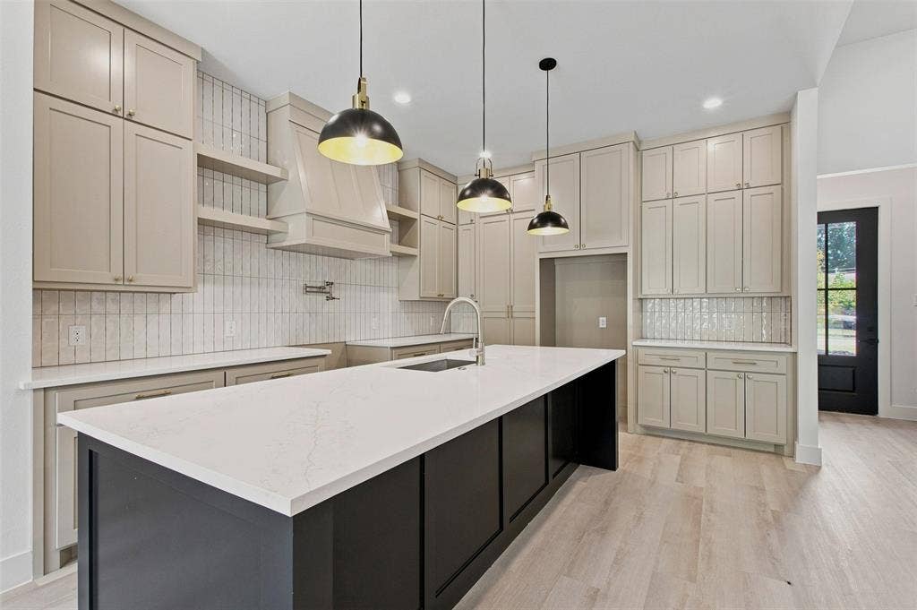 3023 Mallory Drive, Dallas, TX 75216 Spacious kitchen featuring a large island with a light-toned countertop, extensive light-toned cabinetry, light wood-finish flooring, a decorative hood, and a full-height tile backsplash