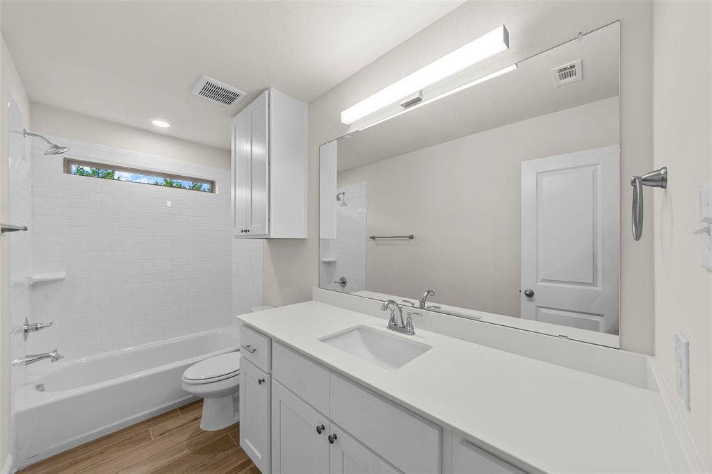 White subway tile tub surround, elongated vanity mirror, white cabinetry, wood-finish flooring, and white solid surface countertop