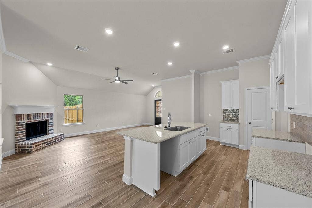 Vaulted ceiling with recessed lighting, wood-finish flooring, brick fireplace with mantel, kitchen island with sink, and white cabinetry