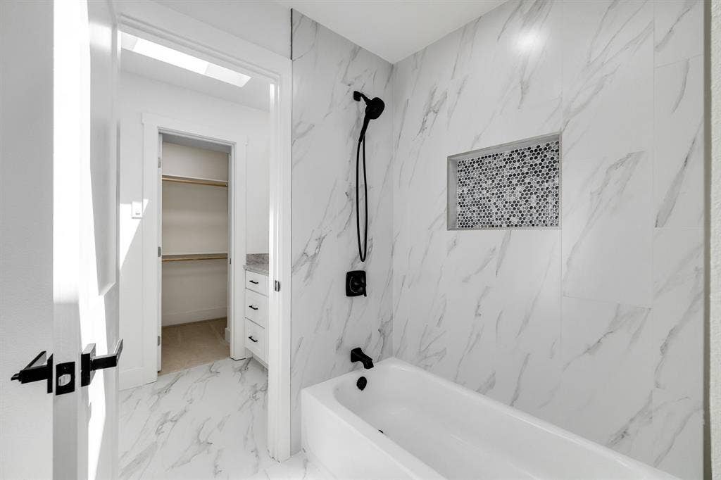 4402 Briargrove Lane, Dallas, TX 75287 4402 Briargrove-Spa-inspired bathroom featuring marble-look tile walls, a white soaking tub with chrome fixtures and handheld showerhead, a recessed niche with mosaic tile accent, and direct access to a walk-in closet
