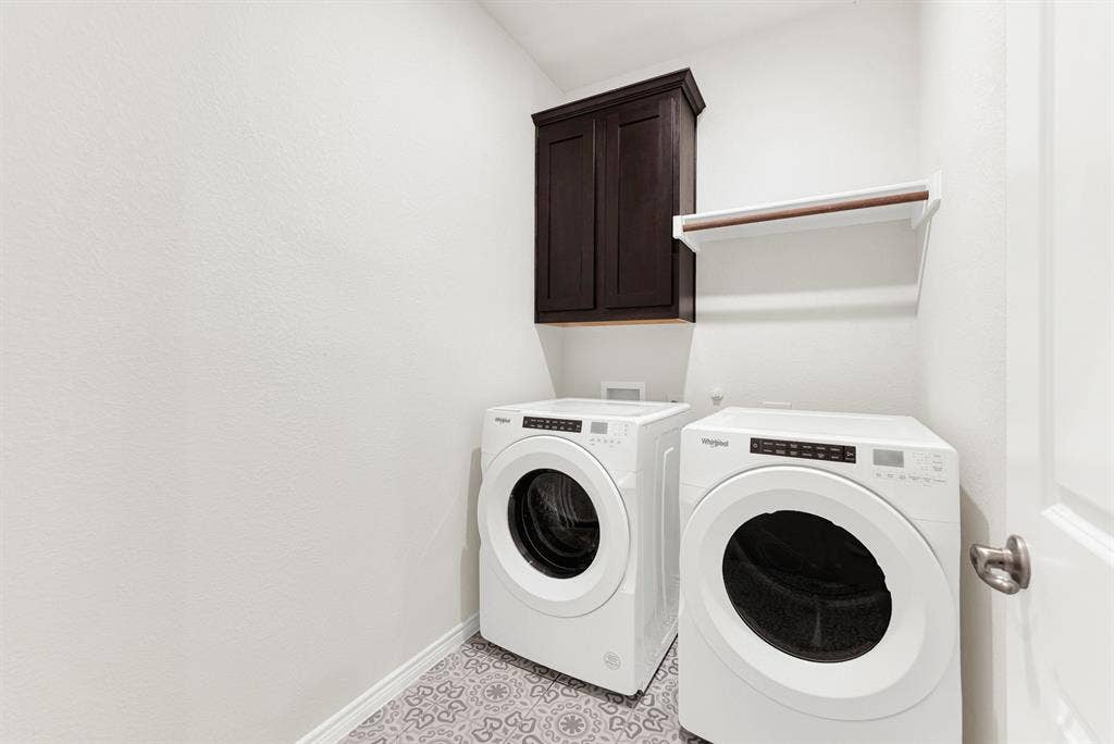 The functional utility room features patterned tile floors, upper cabinetry, and a dedicated hanging rod above modern front-load laundry units. Keep daily chores organized and hassle-free in this clean, stylish, and practical space.