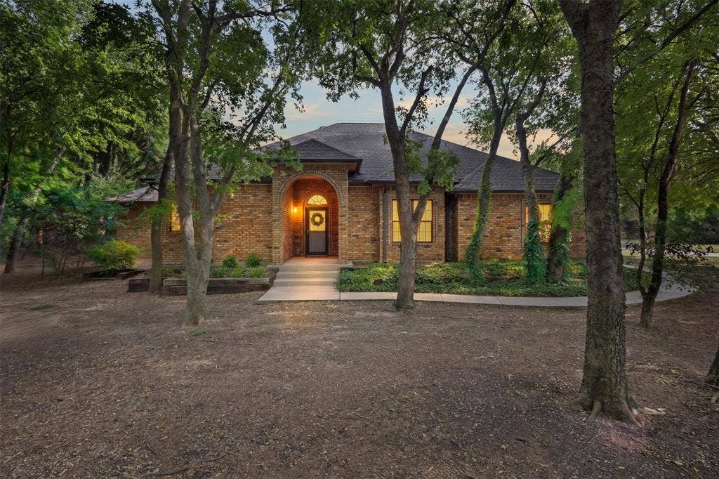 Brick exterior featuring an arched entry, dark shingle roof, concrete walkway, and established trees
