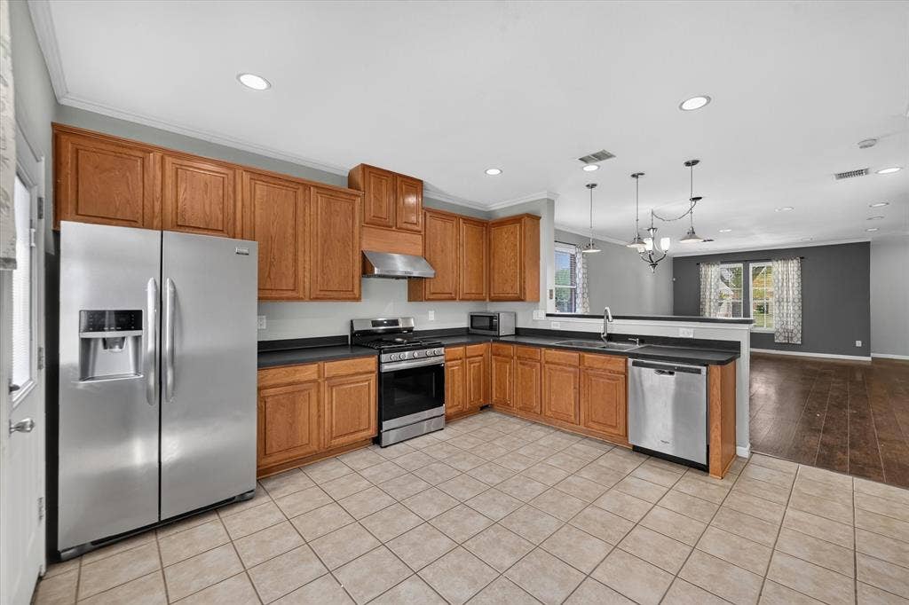 6513 Cartier Court, McKinney, TX 75072 Spacious kitchen featuring warm wood cabinetry, stainless steel appliances including a side-by-side refrigerator and gas range, black countertops, tile flooring, recessed and pendant lighting, and a large island with seating area