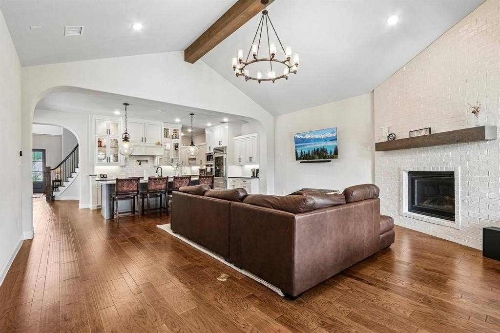 1657 Lilac Lane, Celina, TX 75009 Vaulted ceiling with wood beam, wood-finish flooring, white brick fireplace with mantel, white kitchen cabinetry, and a kitchen island with pendant lighting