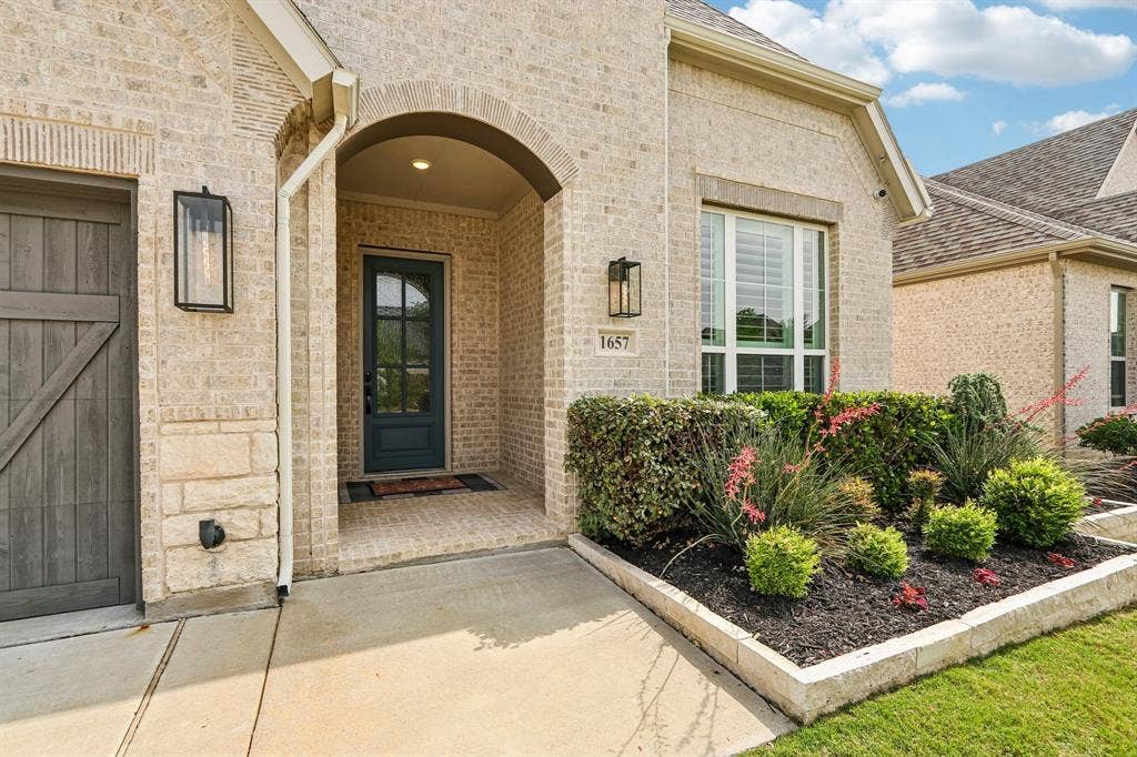 1657 Lilac Lane, Celina, TX 75009 Arched entryway with a dark paneled door, light brick facade, stone accents, and exterior lighting fixtures