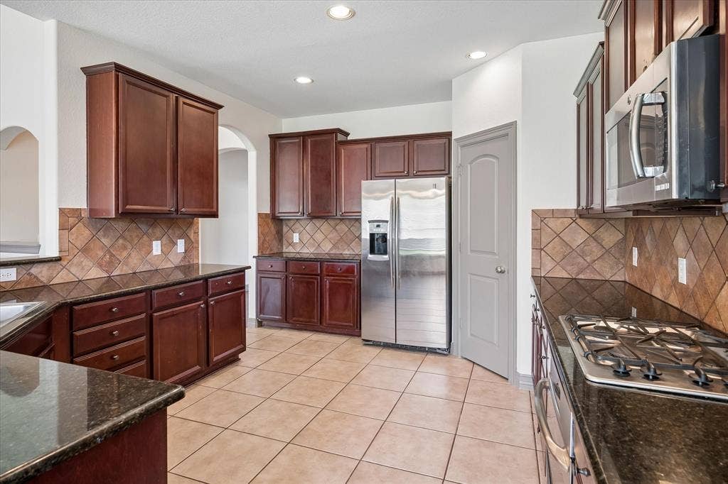 Kitchen featuring dark wood-finish cabinetry, dark countertops, a tiled backsplash, stainless steel appliances, and neutral-toned floor tiling