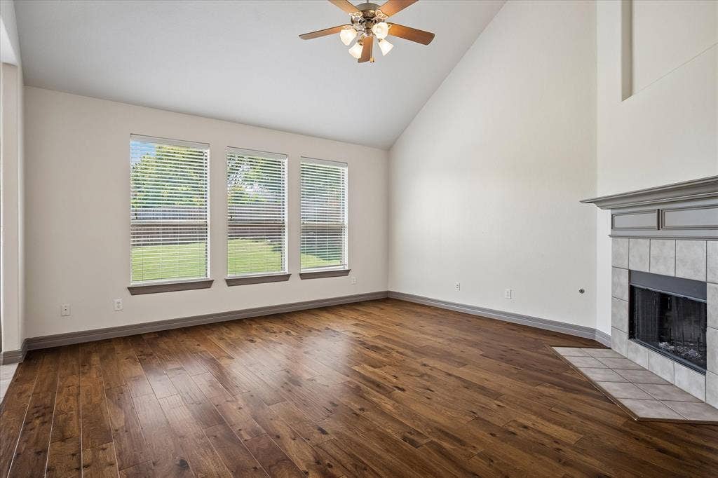 Wood-finish flooring throughout, vaulted ceiling, ceiling fan with integrated lighting, three windows with blinds, and a tiled fireplace with a mantel