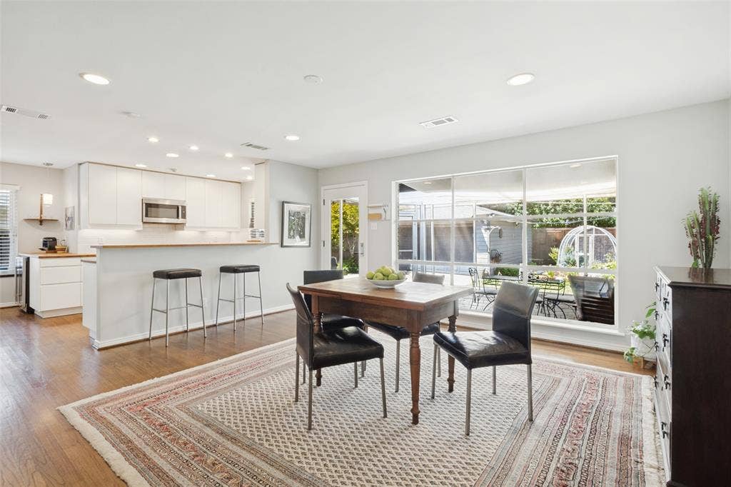 6719 Braeburn Drive, Dallas, TX 75214 Wood-finish flooring throughout, kitchen with white cabinetry and stainless steel microwave, breakfast bar with pendant lighting, recessed ceiling lighting, and large windows overlooking a private outdoor space