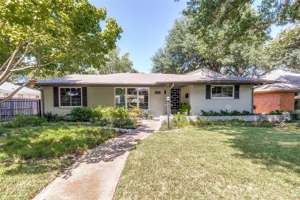 6719 Braeburn Drive, Dallas, TX 75214 Light-colored brick exterior featuring a shingled roof, mature trees, and a concrete pathway