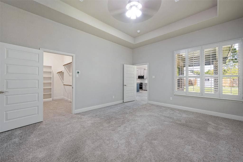 6001 Allison Drive, Midlothian, TX 76065 Recessed tray ceiling with ceiling fan, plush gray carpet, a walk-in closet with built-in shelving, and a large window with plantation shutters are features of the primary bedroom.