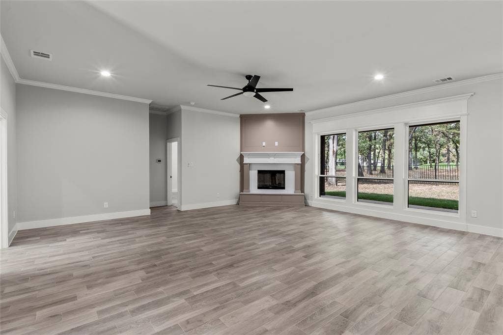 128 Saint Andrews Drive E, Mabank, TX 75156 Spacious room featuring wood-finish flooring, a corner fireplace with white mantle, crown molding, a ceiling fan, and large windows with exterior landscaping