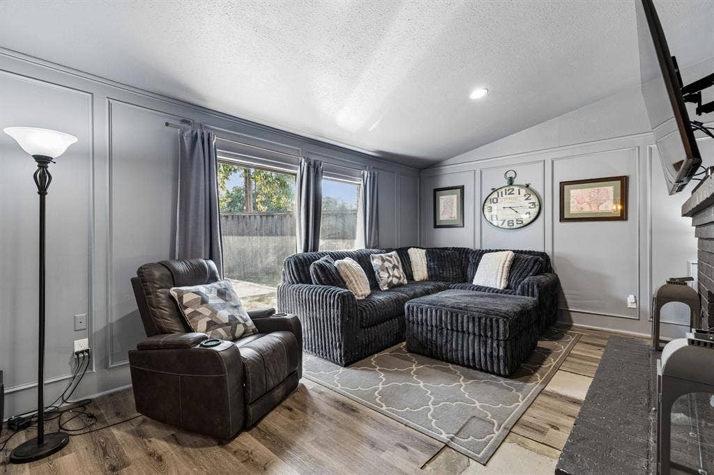 Comfortable living room featuring a sectional configuration with striped upholstery, wood-finish flooring, a geometric area rug, large windows with blue drapery, recessed ceiling lighting, and wall-mounted clock and artwork