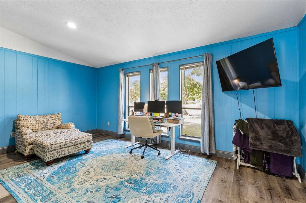 Room with wood-finish flooring, three windows with vertical blinds, and a mounted display