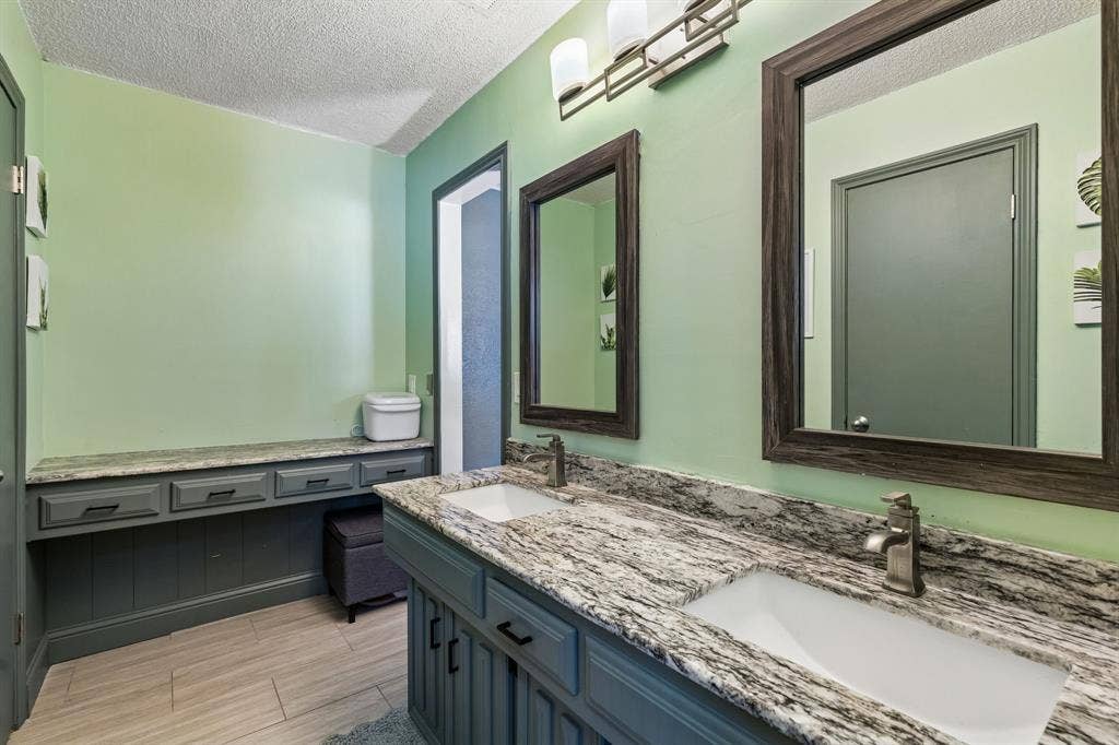 Dual vanity with under-mount sinks, patterned stone countertop, individual framed mirrors, and wood-finish tile flooring