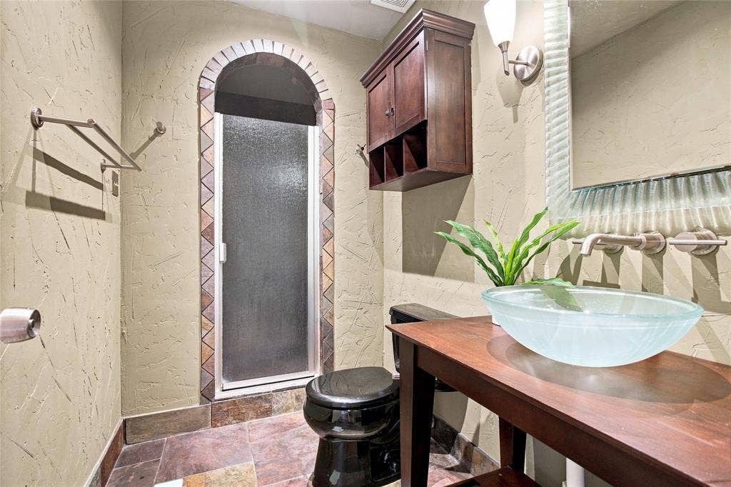 1417 Sun Valley Drive, Garland, TX 75043 Bathroom with wall finish, arched shower entry with decorative tile, glass vessel sink, wall-mounted faucet, and dark wood vanity with cabinet storage