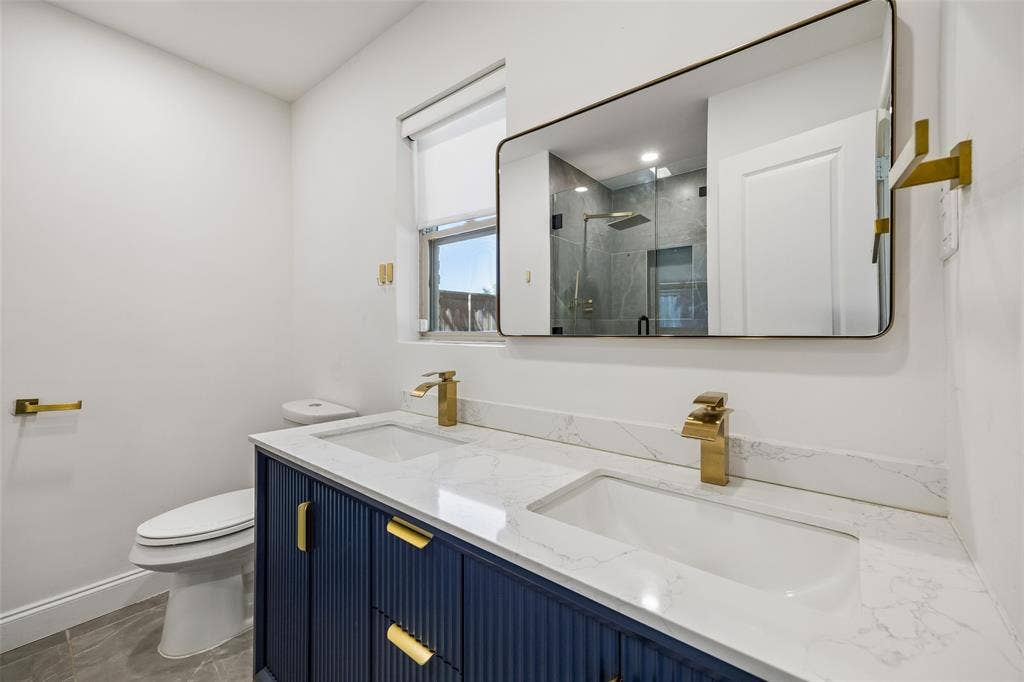 5610 Forest Lane, Dallas, TX 75230 Dual sinks with brass-finish fixtures, a navy vanity with fluted detailing, and a wall-mounted mirror