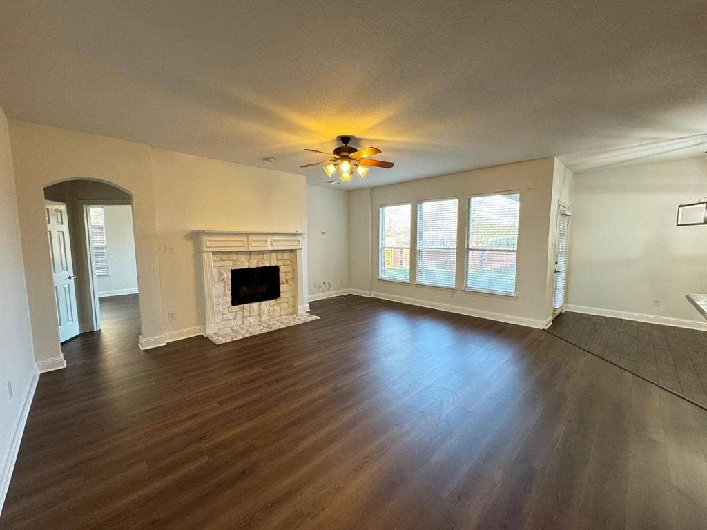 4531 Oak Shores Drive, Plano, TX 75024 Spacious room featuring wood-finish flooring, a stone-surround fireplace with a white mantel, an arched entryway, three large windows, and a ceiling fan