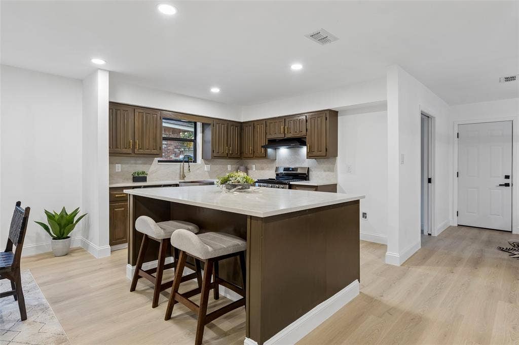 2133 Oak Valley Lane, Dallas, TX 75232 Central kitchen island with a light-toned countertop, wood-finish flooring, recessed lighting, brown cabinetry, and a tile backsplash