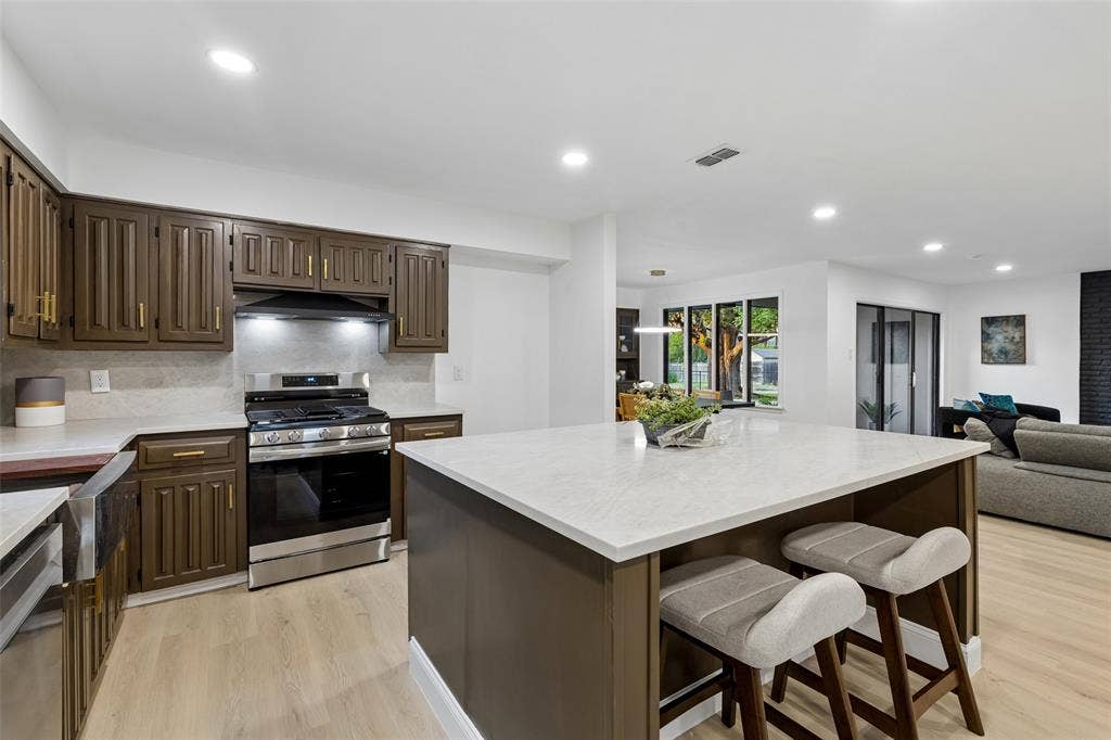 2133 Oak Valley Lane, Dallas, TX 75232 Modern kitchen featuring an expansive island with a light-toned countertop, stainless steel appliances, dark wood-finish cabinetry, and recessed lighting