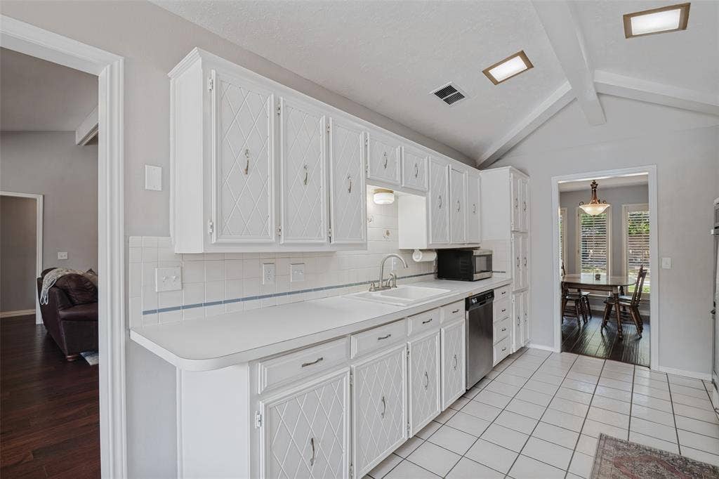 3632 Candelaria Drive, Plano, TX 75023 White cabinetry with diamond pattern detailing, white tile backsplash, vaulted ceiling with exposed beams, stainless steel dishwasher, and tile flooring