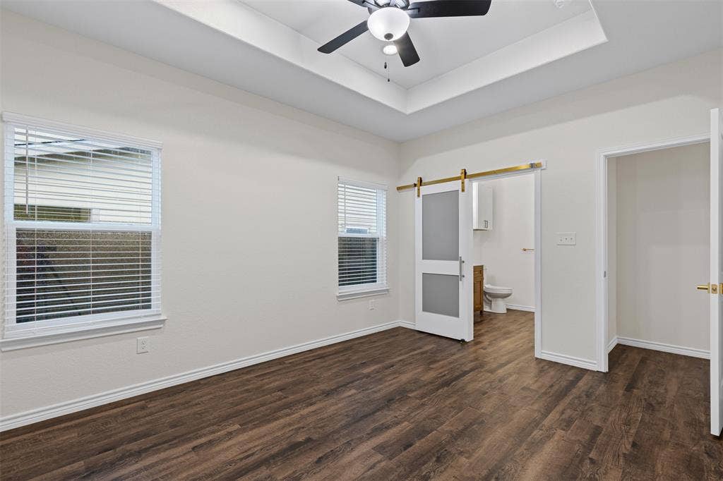 Unfurnished bedroom with a barn door, dark wood finished floors, a raised ceiling, and baseboards