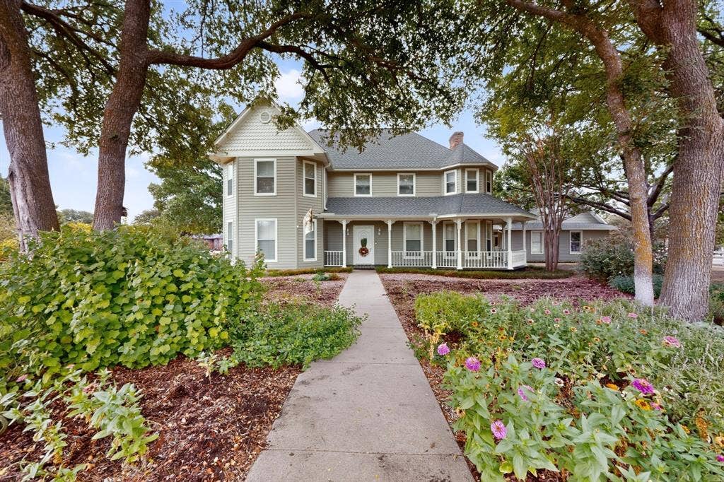 2 Trailridge Drive, Melissa, TX 75454 Two-story exterior with light green siding and white trim, featuring a wrap-around porch with white railings, a prominent gabled dormer, and established landscaping with mature trees and flowering plants