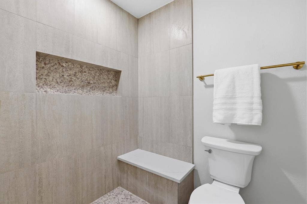 3405 Mill Ridge, Bedford, TX 76021 Tiled shower with a built-in niche featuring decorative stone, an integrated shower bench with a solid surface top, and a white toilet with a brass towel bar