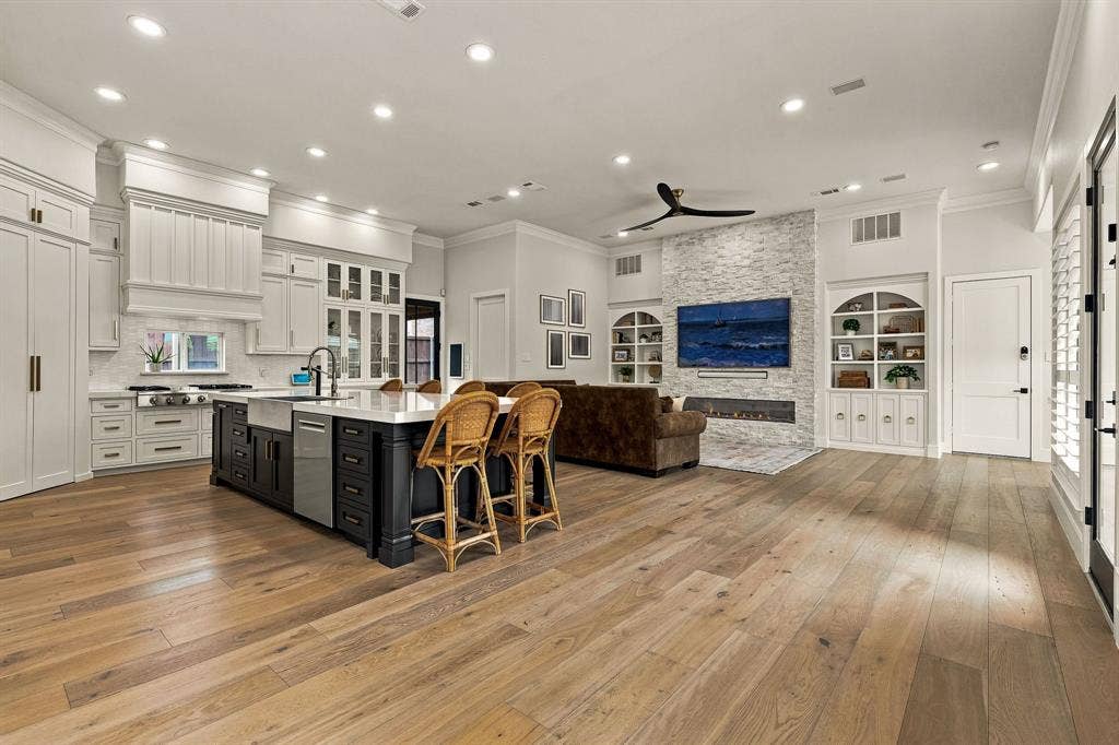 5941 Tiburon Drive, Plano, TX 75093 Wood-finish flooring throughout, kitchen with white upper and lower cabinetry, black island with integrated sink, stone-clad fireplace, and built-in shelving with arched accents