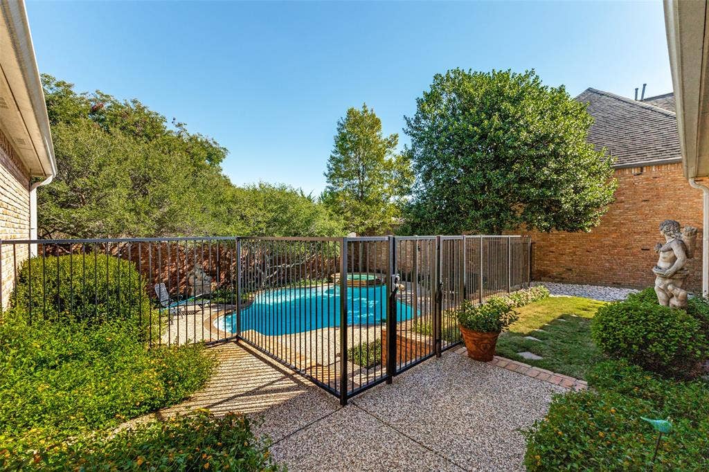12012 Excelsior Way, Dallas, TX 75230 Pebbled concrete patio with a dark metal fence enclosing a pool, surrounded by mature landscaping and a brick-clad exterior