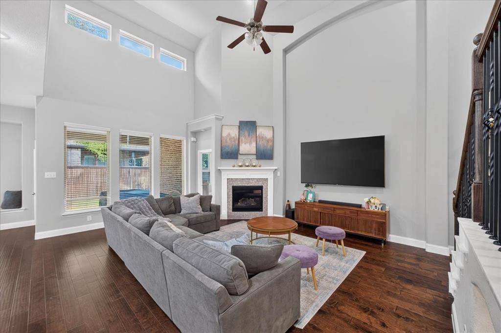 8712 Mccutchins Drive, McKinney, TX 75070 Spacious living area featuring high ceilings, wood-finish flooring, multiple windows, a ceiling fan, and a fireplace with a decorative mantel