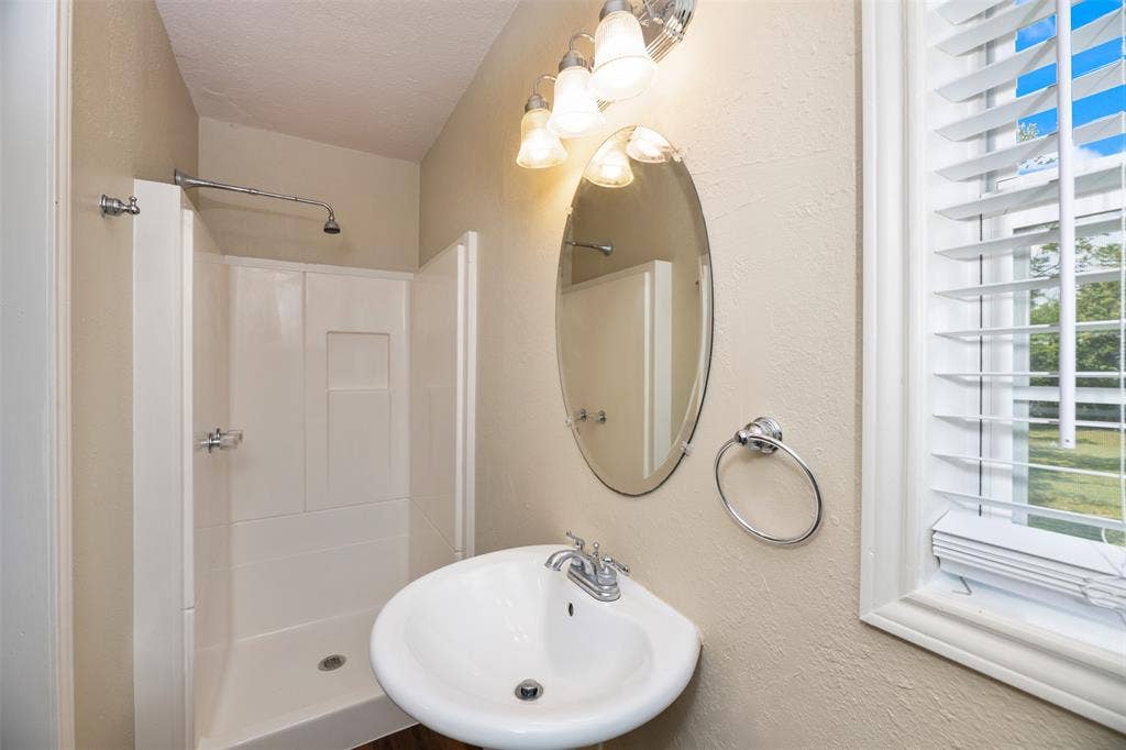 8823 San Benito Way, Dallas, TX 75218 Pedestal sink with chrome faucet, oval mirror, wall-mounted lighting fixture, and a white framed window with horizontal blinds