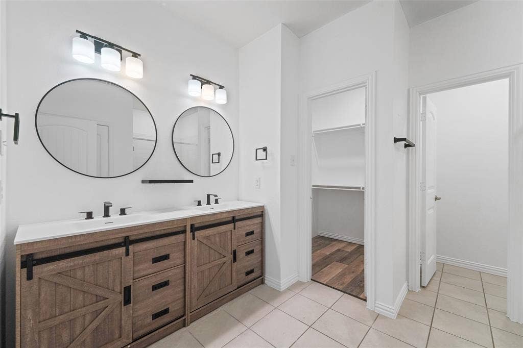 4651 Raleigh Dalton Road, Dallas, TX 75227 Spa-inspired primary bathroom showcasing a custom wood-finish dual vanity with black hardware and metal accents, paired round mirrors with integrated lighting, tile flooring, and a walk-in shower with frameless glass enclosure