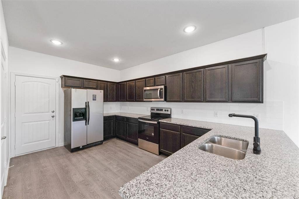 4651 Raleigh Dalton Road, Dallas, TX 75227 Modern kitchen featuring dark wood cabinetry with glass-front upper doors, stainless steel appliances including a refrigerator and range, quartz countertops with waterfall edge, a deep stainless steel sink with matte black pull-down faucet, recessed ceiling lighting, and wood-finish flooring throughout the open layout
