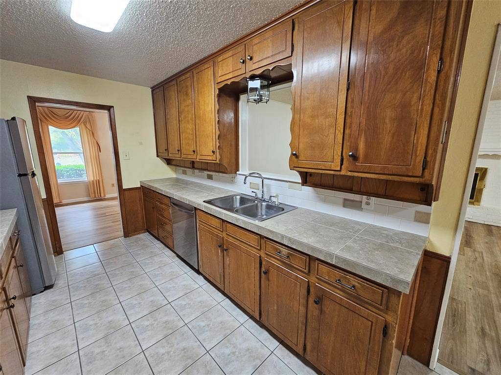 1109 W Redbud Drive, Hurst, TX 76053 Kitchen featuring wood cabinetry, a dual basin stainless steel sink, tile countertops, a white subway tile backsplash, and tile flooring