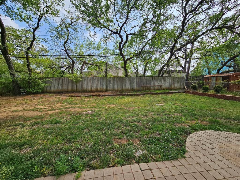 1109 W Redbud Drive, Hurst, TX 76053 Grassed yard with mature trees, perimeter fencing, brick-lined planting beds, and a paver patio