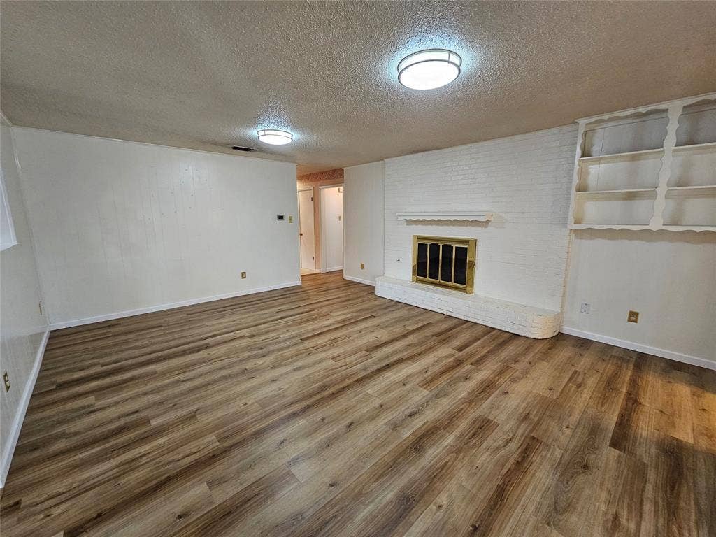 1109 W Redbud Drive, Hurst, TX 76053 Wood-finish flooring, white painted brick fireplace with brass insert, built-in shelving, and two flush-mount ceiling lights
