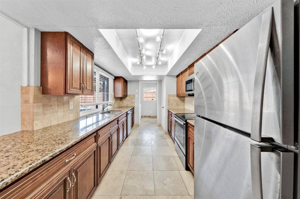 1715 Cherokee Trail, Plano, TX 75023 Galley kitchen featuring wood cabinetry, stone-finish countertops, tile backsplash, stainless steel appliances, and tile flooring